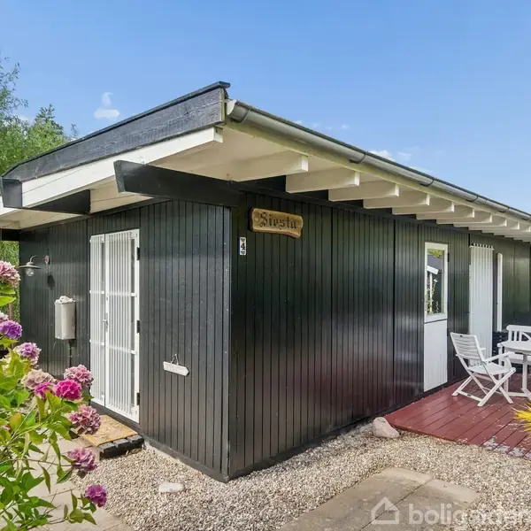 Sort træsommerhus med hvidt tag og døre, omgivet af have med blomster. Et skilt med "Skovstykke" hænger på væggen. En terrasse med hvidt havesæt ses.
