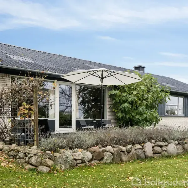 Et enfamiliehus med murstensfacade og tag i mørkegrå tegl, har store vinduer og en terrasse med en parasol. Huset omkranses af en stensætning og grønne omgivelser.