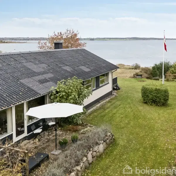 Et enfamiliehus med sort tag og store vinduer står på en grøn græsplæne. En parasol er opstillet i haven. I baggrunden ses havudsigt og en flagstang med Dannebrog.