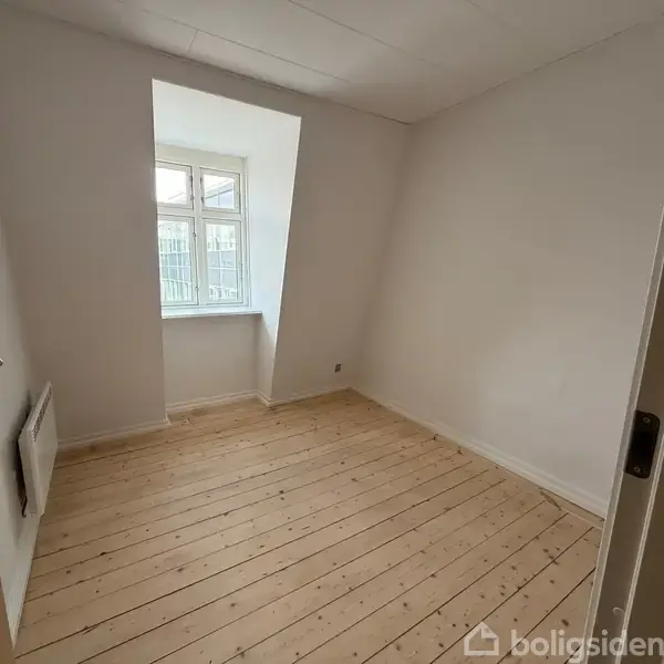 Et lyst rum med trægulv, en skrå væg og et vindue med hvid ramme. En radiator er monteret på venstre væg. Rummet er tomt og nyrenoveret.