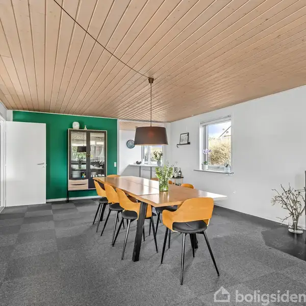 Lang spisebord med seks stole står i en moderne spisestue. Der er en brændeovn i hjørnet, grøn væg i baggrunden, og store vinduer slipper lys ind.