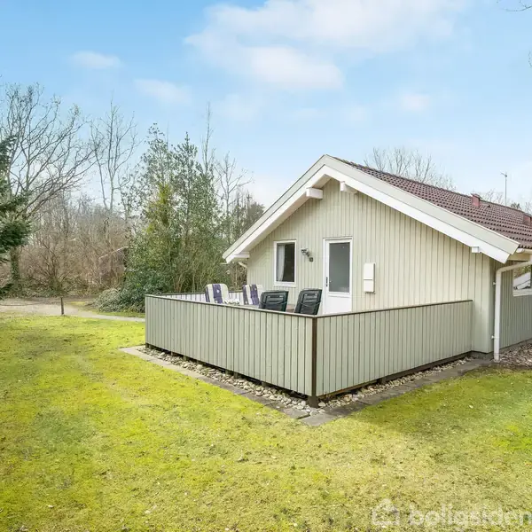 Et lyst træhus står stille omgivet af en grøn have med fyrretræer og nøgne træer i baggrunden. En terrasse med havemøbler er tilknyttet til siden af huset.