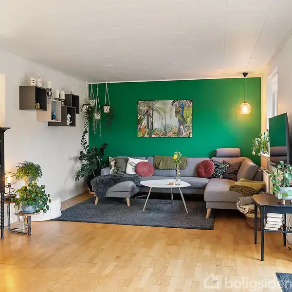 Stue med grå sofa står mod en grøn væg med kunstbillede. Sofaen er pyntet med farverige puder. Rummet har trægulv, planter på hylder og et rundt sofabord i moderne indretning.