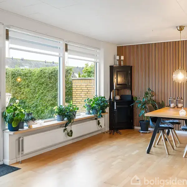 Stue med spisebord og fire stole, trævæg bagved. Store vinduer med udsigt til terrasse og have. Grønne planter på vindueskarmen. Trægulv skaber en lys og indbydende atmosfære.