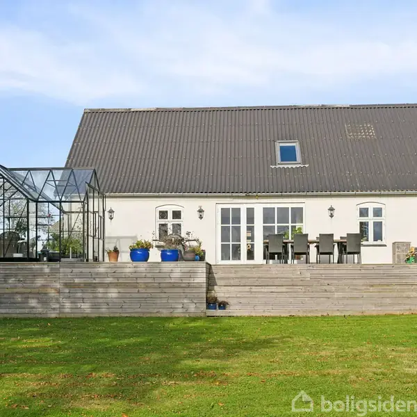 Et hvidt hus med sort tag har en stor træterrasse med havemøbler og drivhus. Terrassen har udsigt til en grøn græsplæne under en lys blå himmel.