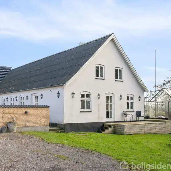 En hvid landlig bygning med sort tag står stille i en have med grøn græsplæne. Ved siden af huset er et drivhus, og foran ligger en terrasse med stuehavemøbler.