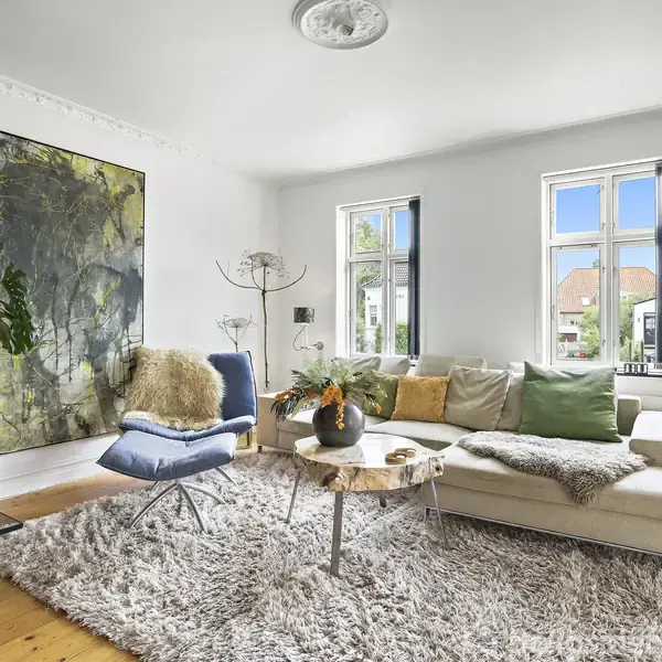 Lænestol står ved siden af en stor, hvid sofa fyldt med puder, placeret på et tæppe i et lyst rum med store vinduer, kunstværk og bogreol i baggrunden.