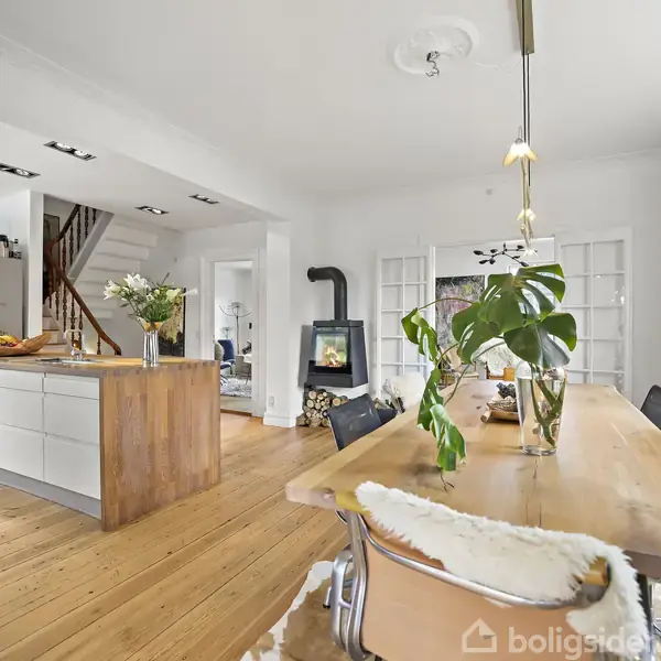 Et træbord med planter står i et moderne køkken-alrum. En køkkenø er dekoreret med frugter og blomster, mens en brændeovn varmelyser rummet med hylder fyldt med bøger.