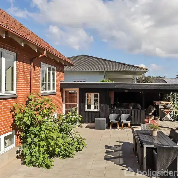 Rødt murstenshus med træafsats, store vinduer. Terrasse har bord og stole, omgivet af grønne planter. Baggrunden viser et anneks med mørkt tag og åben terrasse.