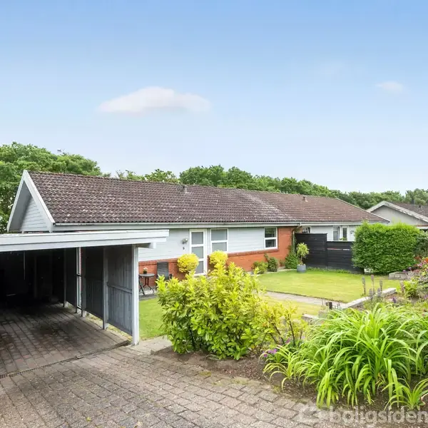 En étplans villa med carport står omgivet af en velplejet have med buske og græsplæne. Villaen har et rødt murstensparti og hvide gavle, beliggende i et grønt område.