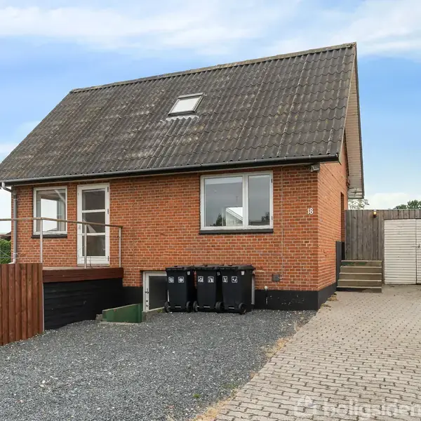 Rødt murstenshus med gråt skråtag; foran står tre affaldsbeholdere. Til højre er en garage med flisebelagt indkørsel. Omkringliggende hegn og grønt buskads.