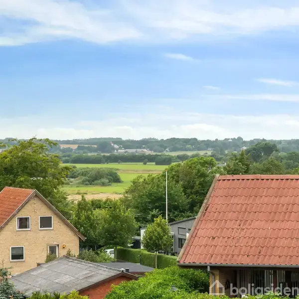 Huse med røde tegltage står i en grøn landsby. I baggrunden er der åbne marker og skove under en klar blå himmel.