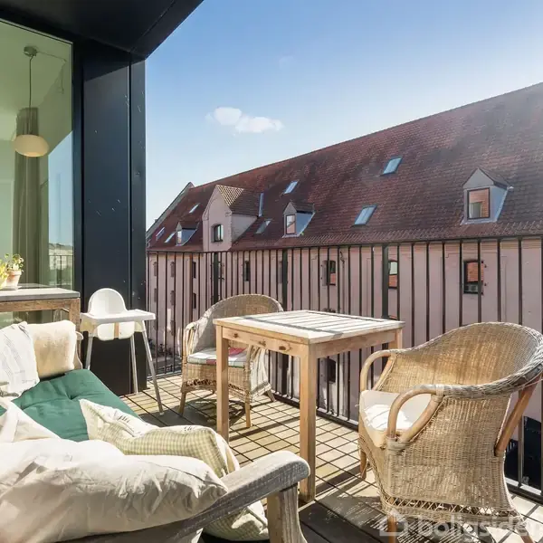 En balkon med møbler, herunder en sofa med puder og to kurvestole, står i sollys. Der er et bord mellem stolene. Baggrunden viser et rødt tegltag på en bygning.