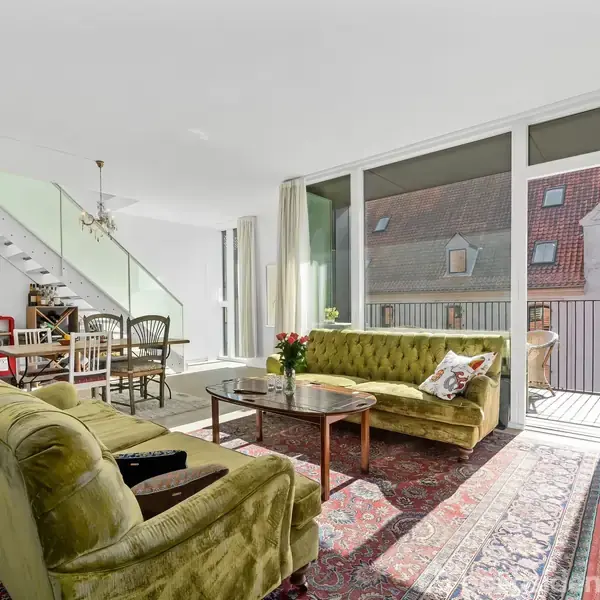 Grønne sofaer står i stue med gulvtæppe, mens daglys strømmer ind gennem store vinduer. En trappe i baggrunden fører til en anden etage. Ingen tekst i billedet.