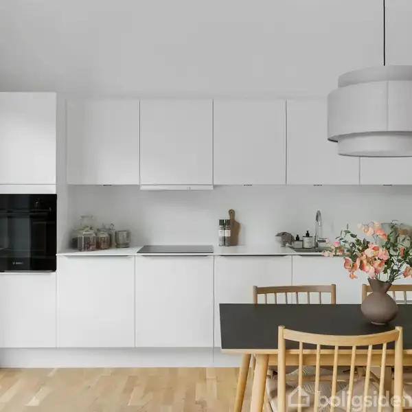 Et moderne køkken med hvide skabe og indbygget ovn. Et spisebord med træstole er dekoreret med en vase blomster. Rummet er lyst med trægulv og stor lampe.