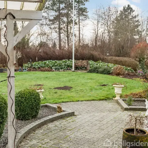 En grøn græsplæne med en hvid statue og dekorative krukker i en velplejet have. Omkring plænen er der buske, træer og en brolagt terrasse.