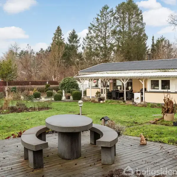 Rundt havebord med tre bænke står på træterrasse. I baggrunden ses en stor græsplæne med buske og træer. Et lille hus med veranda er synligt til højre.
