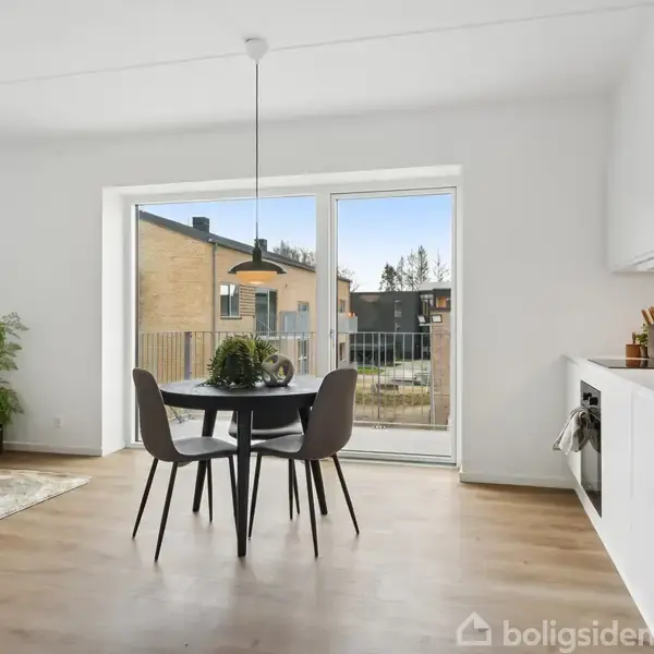 Spisebord med fire stole står foran store vinduer med udsigt til bygninger udenfor. Køkken til højre med hvide skabe og planter ved siden af sofa i hjørnet.