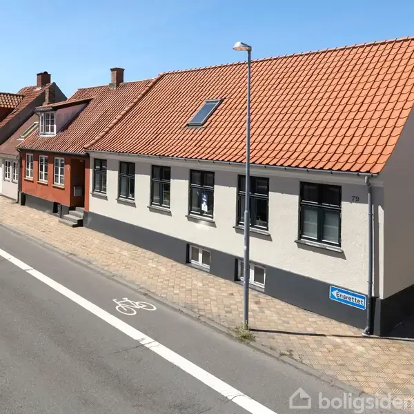Et rækkehus med hvid facade og rødt tegltag står langs en vej med cykelsti. Bygningen har flere vinduer og en gadelampe foran. Tæt på andre lignende huse.