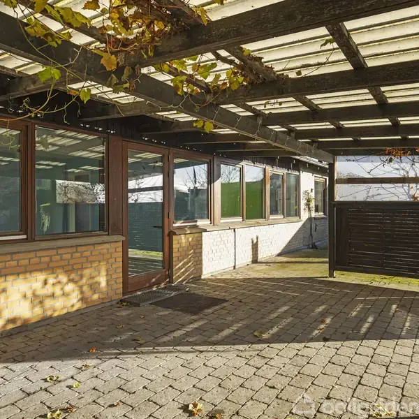 En carport er dækket af et gennemsigtigt tag med slyngplanter ovenpå. Murstensvægge omkranser området med store vinduer. Flisebelagt gulv danner bunden i et solbeskinnet miljø.