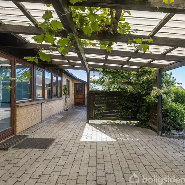 Overdækket terrasse med hvidt træloft og planter, hvor der vokser grønt løv. Murstenshus med store vinduer til venstre, brolagt gulv og have i baggrunden.