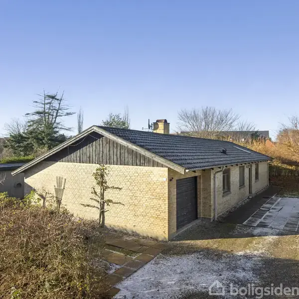 En enkeltliggende villa med fladt tag og gule mursten står stille i en have omkranset af buske og træer. En stenbelagt indkørsel fører til garagen ved siden af.