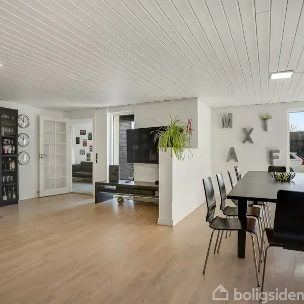 En stue med spisebord og otte stole, omgivet af bøger på en hylde, tre vægure og en fladskærm. Hvide vægge og trælaminatgulve skaber en moderne atmosfære.