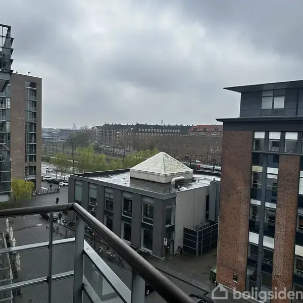 Balkon med metalgelænder har udsigt over en by med moderne bygninger. Det er overskyet, og man kan se træer og veje i baggrunden.