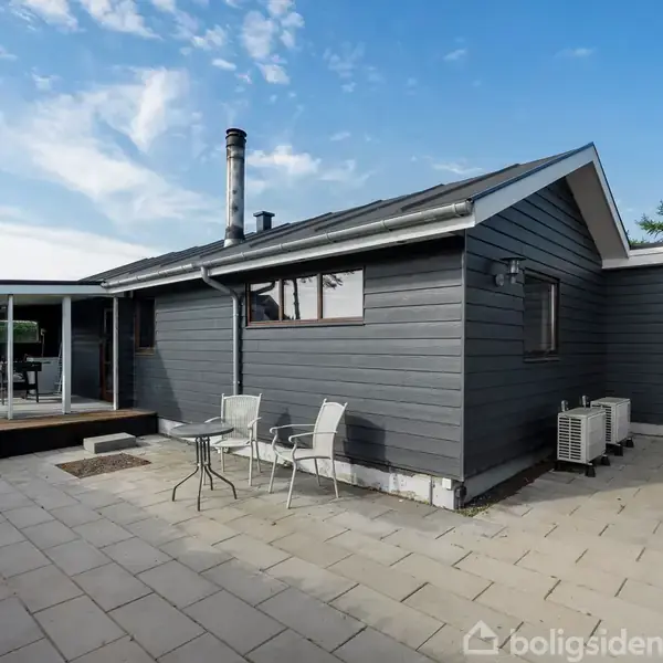Gråt hus står stille på en rummelig terrasse med fliser. To stole og et bord foran huset. Omkringliggende have og blå himmel med få skyer i baggrunden.