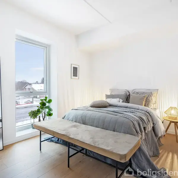 Seng dækket med gråt sengetøj står i et lyst soveværelse. En bænk foran sengen, lampe på natbord og en plante ved vinduet. Store spejle op ad væggen.