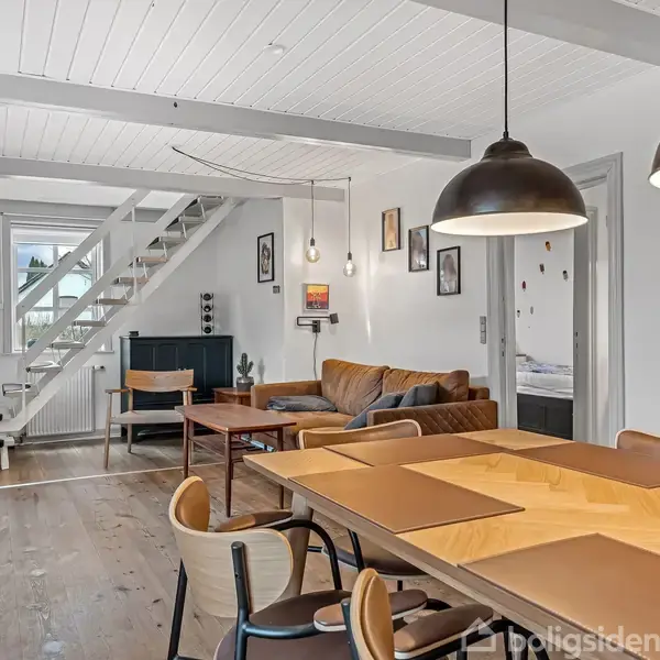 Kontorstol foran et skrivebord med computer og skuffemøbel i et rum med skråvæg og lille vindue. Der er tæppe på gulvet, og en kommode med printer står til højre.