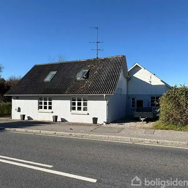 Et hvidt hus med mørkt tag står stille ved siden af en vej. Omgivet af buske og en klar blå himmel i baggrunden.