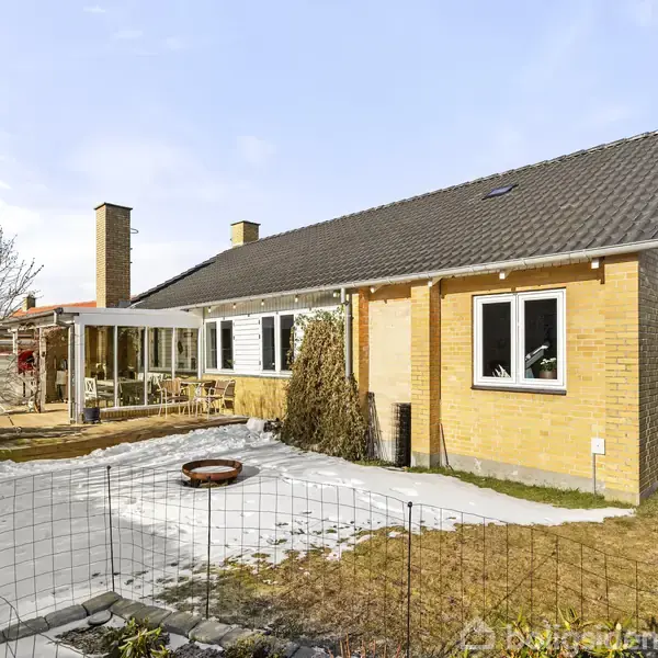 Gult murstenshus med mørkt tag står stille i en have, omgivet af et trådhegn. En skovl ligger ved siden af en sneklædt græsplæne. Der er en terrasse i baggrunden.