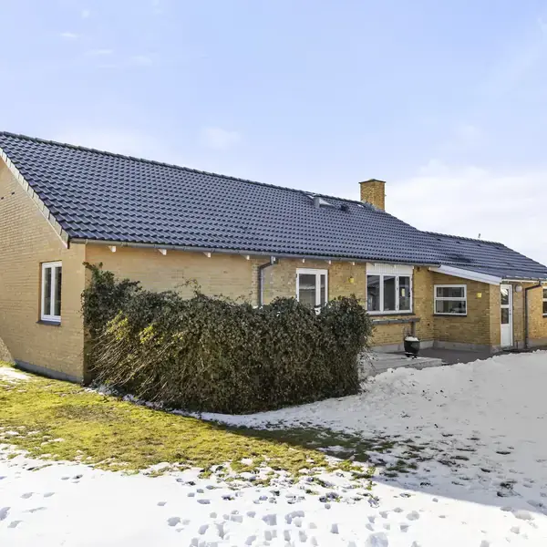 Et gulstenshus med mørkt tag står i en have dækket delvist af sne. En garage er til højre i billedet, mens græsset er synligt til venstre.