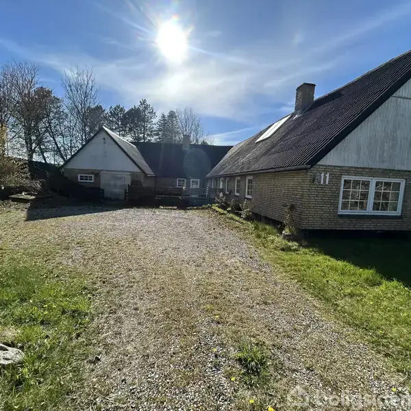 Hus udført i lyse mursten står under en klar, solrig himmel. Foran huset er der en grusvej omgivet af græsplæner. Omkring er der spredte træer og buske.