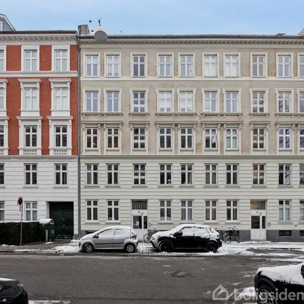 En række historiske bygninger med flere etager står side om side. Foran bygningerne holder biler parkeret på en sneklædt vej. Træer er synlige i baggrunden.