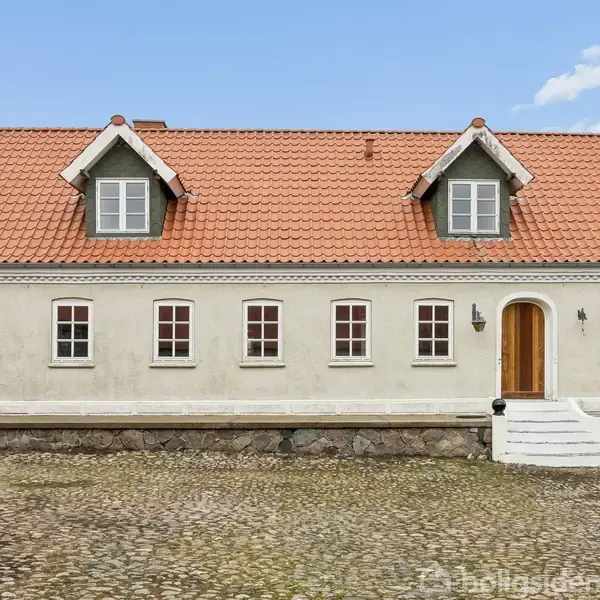 Køkken uden møbler med træbordplader og -hylder, hvide skabe og ovn. Store vinduer giver naturligt lys, lysekroner hænger fra loftet, og mørkt trægulv dækker hele rummet.