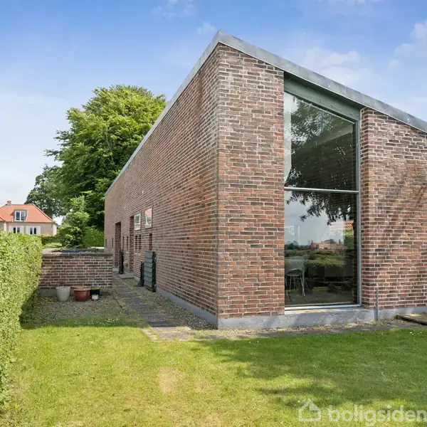 Floden flyder stille gennem det grønne landskab, omgivet af træer og et nabolag med huse. I baggrunden ses flere bygninger og en vid horisont.