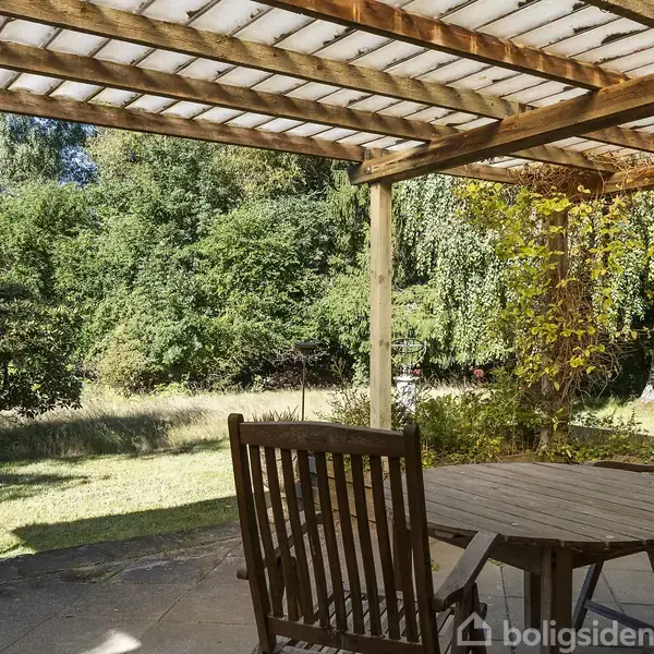 Terrassebord med stole står under en åben pergola. En grøn have med tæt bevoksning omkranser området, hvilket skaber en hyggelig og skyggefuld atmosfære i et naturpræget miljø.