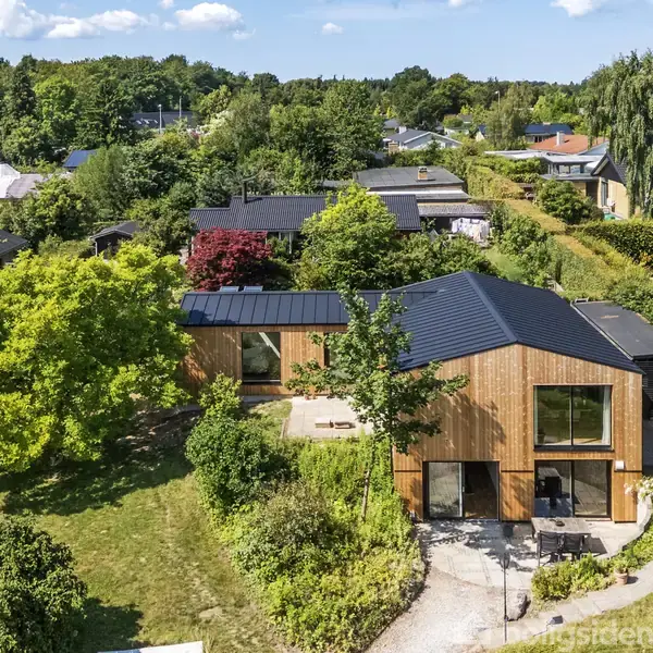 Moderne træhus med sorte tagplader ligger omgivet af frodig have med forskellige træer og buske. Huset har store vinduespartier og en terrasse. Baggrund viser flere huse og grønne områder.