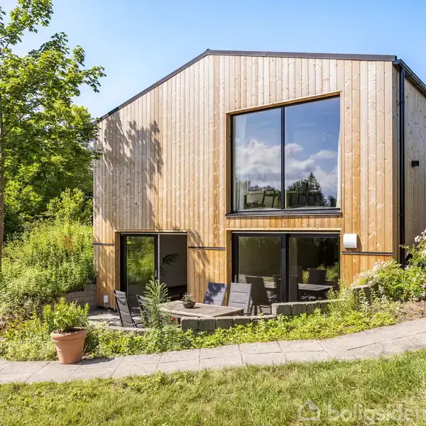 Et moderne træhus med store vinduer står i en grønt område. En terrasse foran huset har havemøbler omgivet af tæt beplantning og velplejet græsplæne.