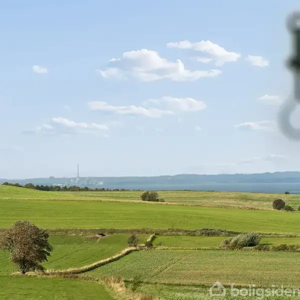 En krog ses i forgrunden. Udsigt over grønne marker strækker sig mod horisonten under en blå himmel med lette skyer. I baggrunden ses en fjern radiomast.