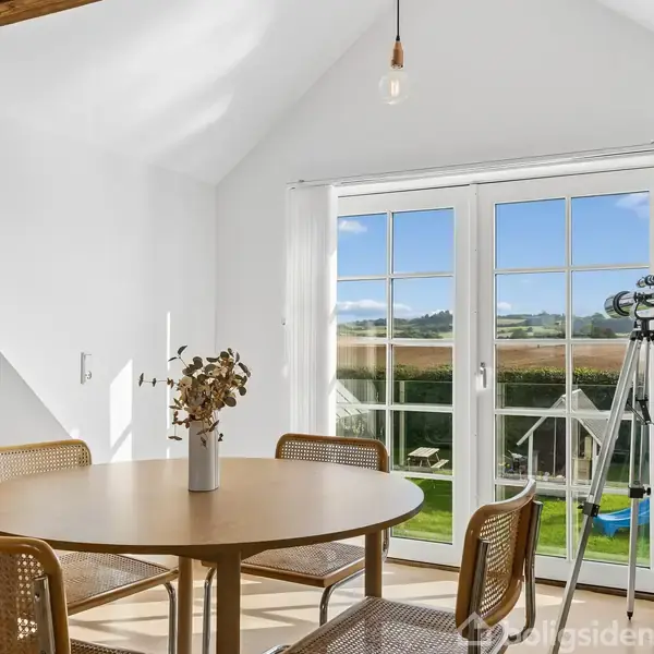 Rundt bord omgivet af fire stole står i lys stue. En vase med blomster er på bordet, mens en kikkert på stativ peger mod vinduet, der viser udsigt til have og landskab.