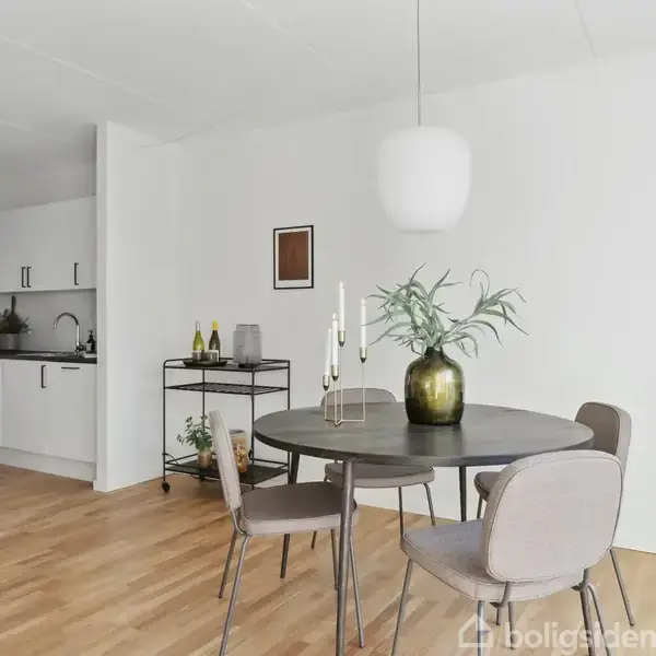 Et rundt spisebord med fire stole står i en moderne spisestue. En grøn vase med blade og lysestage pryder bordet, mens et åbent køkkenområde ses i baggrunden.