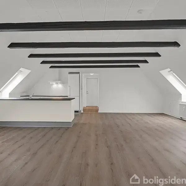 Loftet har et lyst rum med synlige mørke bjælker. Rummet indeholder et åbent køkken med en ø, placeret mellem to skråvinduer, og har et lyst trægulv.