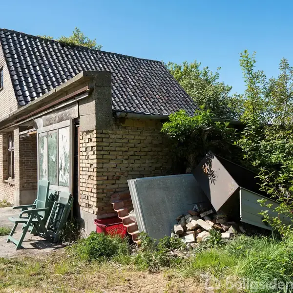 Et gammelt murstenshus med sort tegltag står forladt. Multiple genstande, herunder havestole og affald, er stablet foran huset. Omkringliggende vegetation vokser tæt.