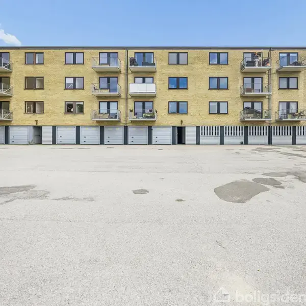 En tre-etagers gul murstensbygning har balkoner, mens garager ligger i stueetagen. Foran er der en stor asfalteret plads. Til højre ses affaldscontainere i forskellige farver.