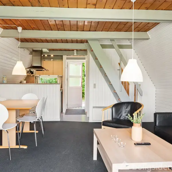 Spisestuens bord og stole står stille i et lyst, åbent rum med hvidmalede vægge og trælloftet. Køkken i baggrunden og en trappe leder til en anden etage.
