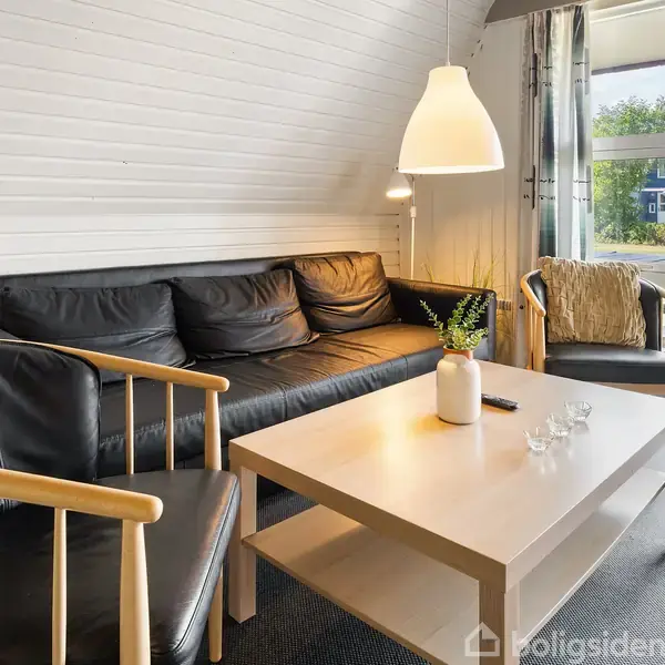 Sorte læderstole og en sofa står omkring et lyst sofabord med en vase. Et stort vindue åbner ud til en terrasse med udsigt over en have og nabohuse.