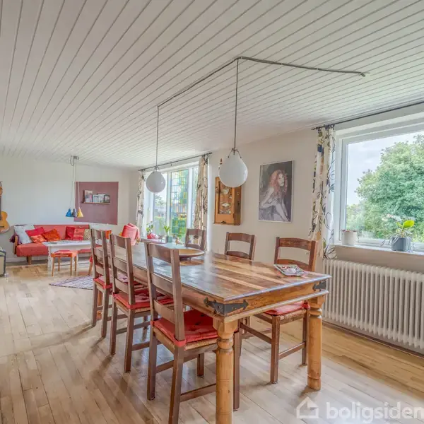 Spisebord med stole står i en stue. Et maleri hænger på væggen. Sofaområde og guitar ses i baggrunden, mens vinduer med udsigt til have er på højre side.